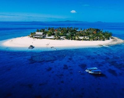Small tropical island with white sandy beaches, lush green vegetation, and several buildings, surrounded by clear blue water with two boats nearby, under a bright blue sky with distant landmasses in the background