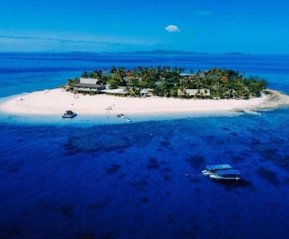 Small tropical island with white sandy beaches, lush green vegetation, and several buildings, surrounded by clear blue water with two boats nearby, under a bright blue sky with distant landmasses in the background