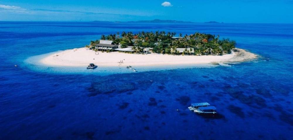 Small tropical island with white sandy beaches, lush green vegetation, and several buildings, surrounded by clear blue water with two boats nearby, under a bright blue sky with distant landmasses in the background