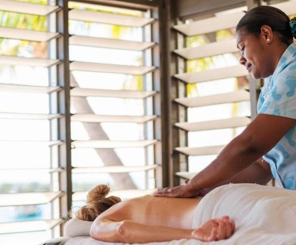 A person receiving a back massage while lying face down on a massage table covered with a white sheet. The massage is being given by someone in a light blue patterned shirt, in a bright room with large windows and horizontal blinds, suggesting a spa or wellness setting
