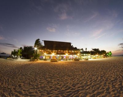 Beachfront scene at dusk featuring a well-lit building, likely a resort or restaurant, surrounded by palm trees on a sandy beach. The sky shows hues of blue and pink as it transitions from day to night