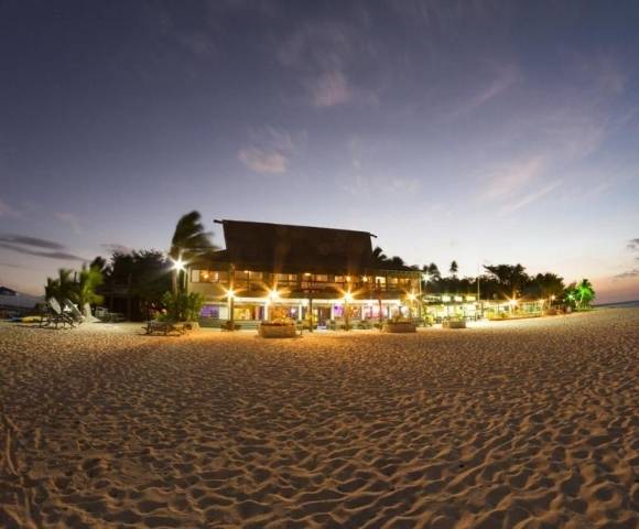Beachfront scene at dusk featuring a well-lit building, likely a resort or restaurant, surrounded by palm trees on a sandy beach. The sky shows hues of blue and pink as it transitions from day to night