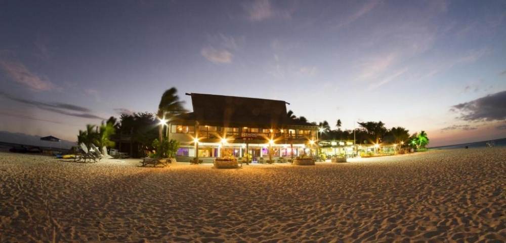 Beachfront scene at dusk featuring a well-lit building, likely a resort or restaurant, surrounded by palm trees on a sandy beach. The sky shows hues of blue and pink as it transitions from day to night