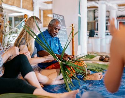 Group of people seated on the floor engaging in a weaving activity using green palm leaves, with one person demonstrating the technique while others observe and participate in a communal setting