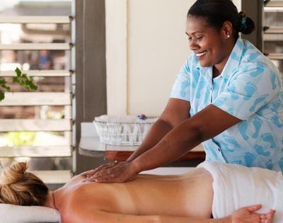 Person receiving a back massage in a spa setting, lying face down on a towel-covered table, with the masseuse wearing a patterned light blue shirt; background features windows with blinds and visible greenery