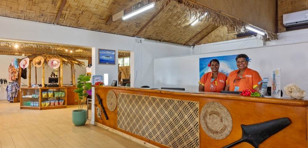 Reception area with two people behind a wooden counter featuring woven patterns and traditional artifacts, under a thatched ceiling with fluorescent lights; background includes a shop with shelves displaying various products in a tropical resort setting
