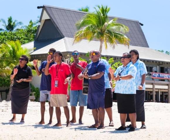 Group of people standing on sandy ground in front of a thatched-roof building with palm trees, some holding musical instruments, in a tropical outdoor setting
