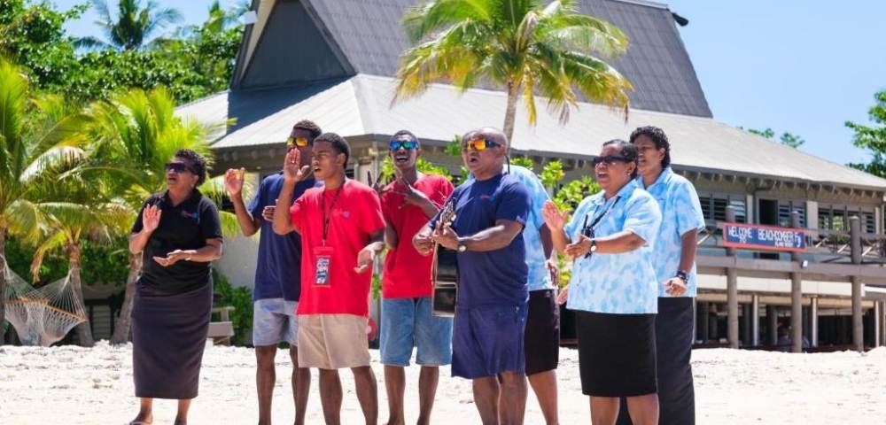 Group of people standing on sandy ground in front of a thatched-roof building with palm trees, some holding musical instruments, in a tropical outdoor setting