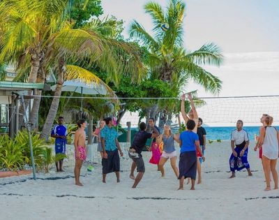 Group of people playing volleyball on a sandy beach with palm trees and ocean in the background, dressed in casual beachwear and actively engaged in the game