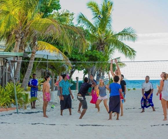 Group of people playing volleyball on a sandy beach with palm trees and ocean in the background, dressed in casual beachwear and actively engaged in the game