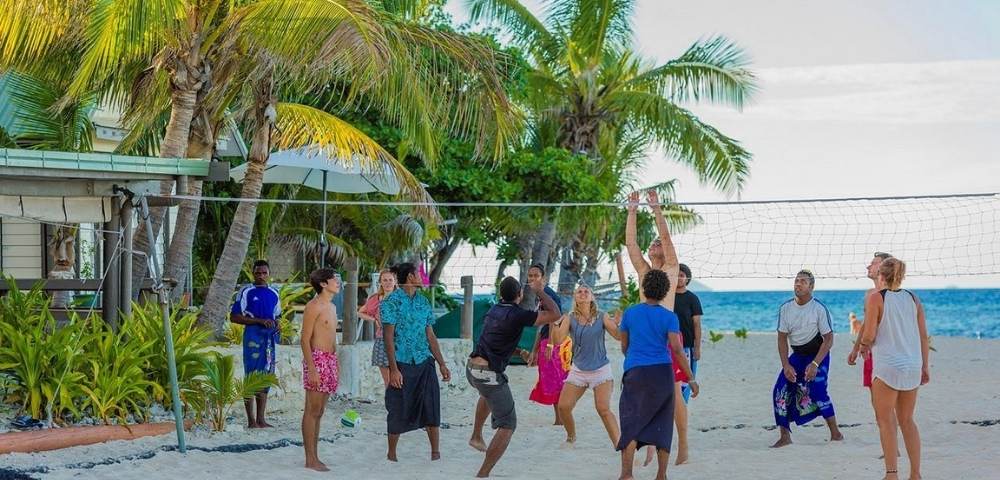 Group of people playing volleyball on a sandy beach with palm trees and ocean in the background, dressed in casual beachwear and actively engaged in the game
