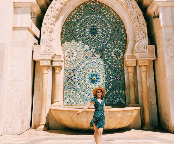 A person stands in front of a beautifully tiled Moroccan fountain adorned with intricate geometric patterns.