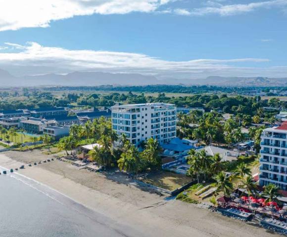 Aerial view of a coastal area with several buildings, dense tree coverage, and the ocean in the background.