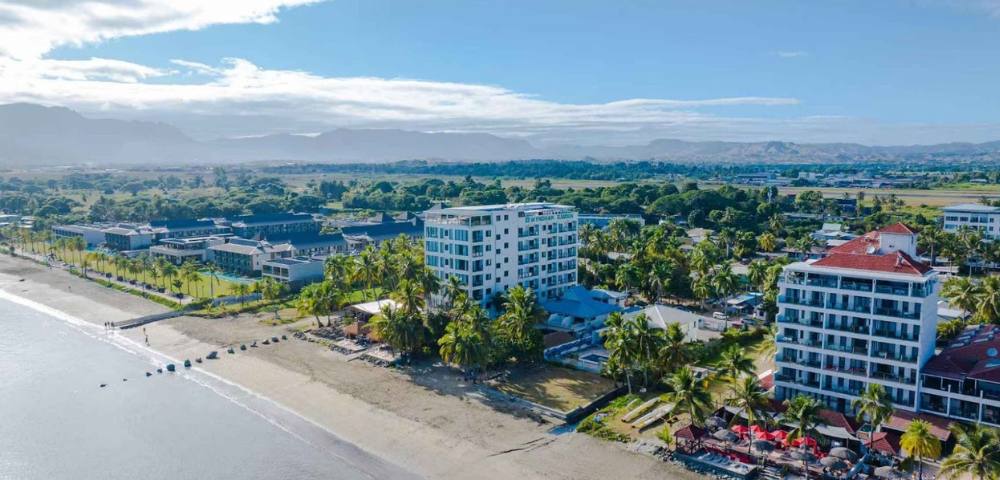Aerial view of a coastal area with several buildings, dense tree coverage, and the ocean in the background.