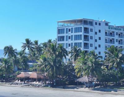 A multi-story building close to the shore, surrounded by trees and overlooking a body of water.
