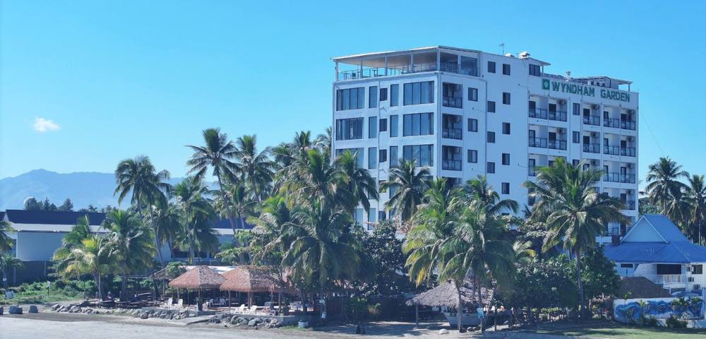 A multi-story building close to the shore, surrounded by trees and overlooking a body of water.