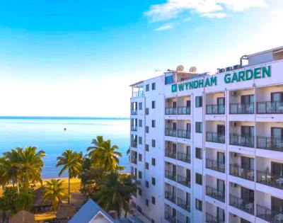 A multi-story white building with balconies, surrounded by lush greenery and overlooking a calm body of water.