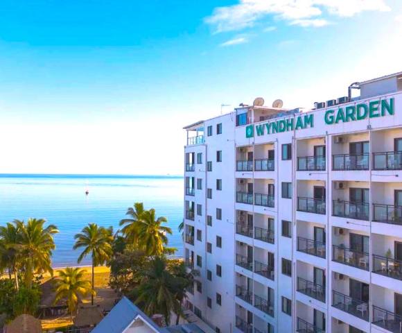 A multi-story white building with balconies, surrounded by lush greenery and overlooking a calm body of water.