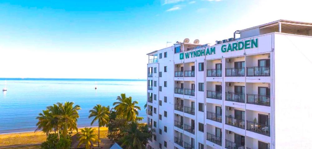 A multi-story white building with balconies, surrounded by lush greenery and overlooking a calm body of water.