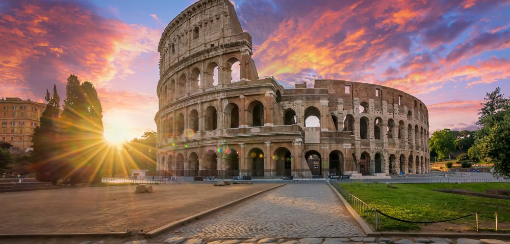 The Colosseum in Rome, Italy, captured at sunset with a vibrant sky of orange, pink, and purple hues. A cobblestone path leads toward the ancient amphitheater, which features multiple arches and levels. The sun creates a starburst effect as it sets behind trees on the left, casting warm light across the scene.