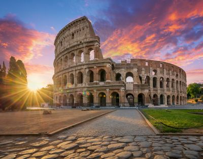 The Colosseum in Rome, Italy, captured at sunset with a vibrant sky of orange, pink, and purple hues. A cobblestone path leads toward the ancient amphitheater, which features multiple arches and levels. The sun creates a starburst effect as it sets behind trees on the left, casting warm light across the scene.