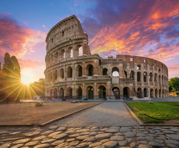 The Colosseum in Rome, Italy, captured at sunset with a vibrant sky of orange, pink, and purple hues. A cobblestone path leads toward the ancient amphitheater, which features multiple arches and levels. The sun creates a starburst effect as it sets behind trees on the left, casting warm light across the scene.