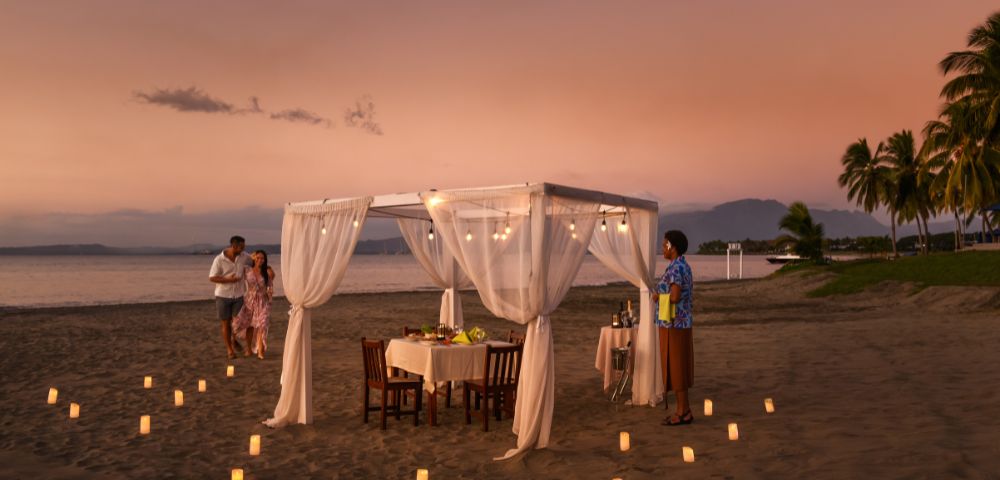 Romantic beachfront dining table with white canopy and sunset view.