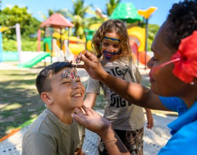 Person applying face paint to a child at an outdoor playground with colorful slides and climbing structures in the background.