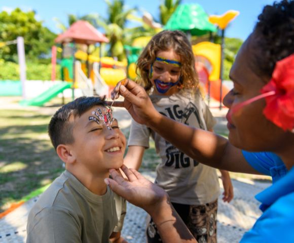 Person applying face paint to a child at an outdoor playground with colorful slides and climbing structures in the background.