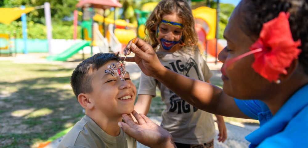 Person applying face paint to a child at an outdoor playground with colorful slides and climbing structures in the background.