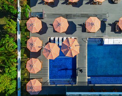 Aerial view of a poolside area with multiple orange-and-white striped umbrellas arranged around a small pool and a larger swimming pool, surrounded by greenery.