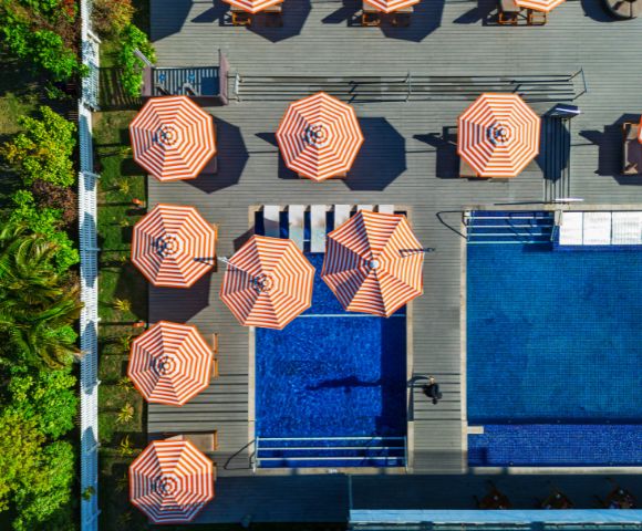 Aerial view of a poolside area with multiple orange-and-white striped umbrellas arranged around a small pool and a larger swimming pool, surrounded by greenery.