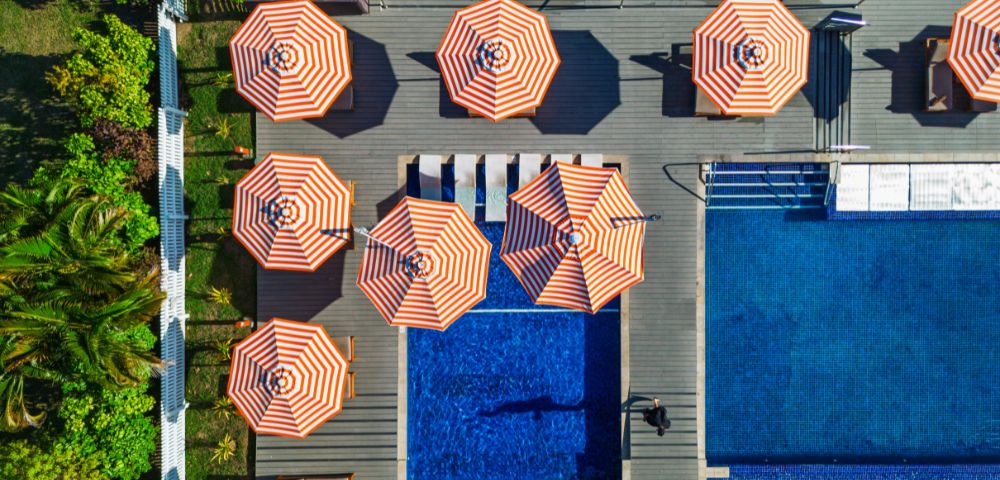Aerial view of a poolside area with multiple orange-and-white striped umbrellas arranged around a small pool and a larger swimming pool, surrounded by greenery.