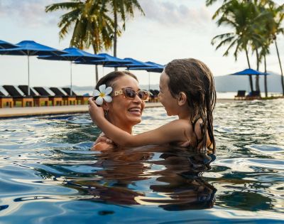 Parent holding a child in a pool with palm trees in the background.