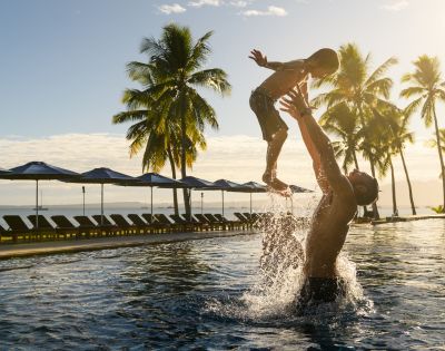 People playing in a resort pool surrounded by palm trees and ocean view.