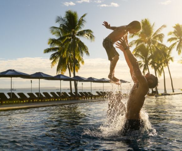 People playing in a resort pool surrounded by palm trees and ocean view.