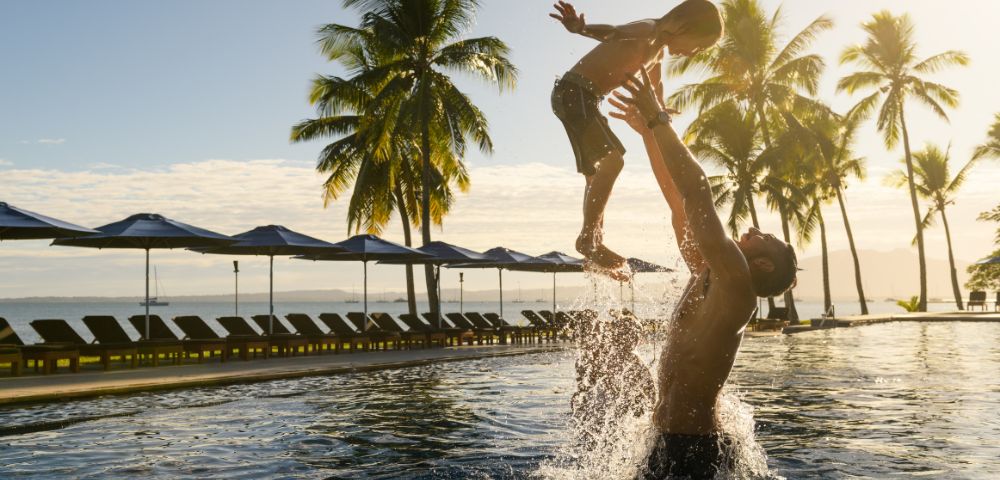 People playing in a resort pool surrounded by palm trees and ocean view.
