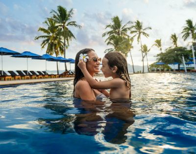 Person holding a child in a pool with palm trees and beach in the background.