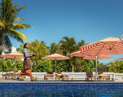 Resort staff walking along a poolside deck carrying a tray, with orange-and-white striped umbrellas, lounge chairs, palm trees, and a clear blue sky in the background.