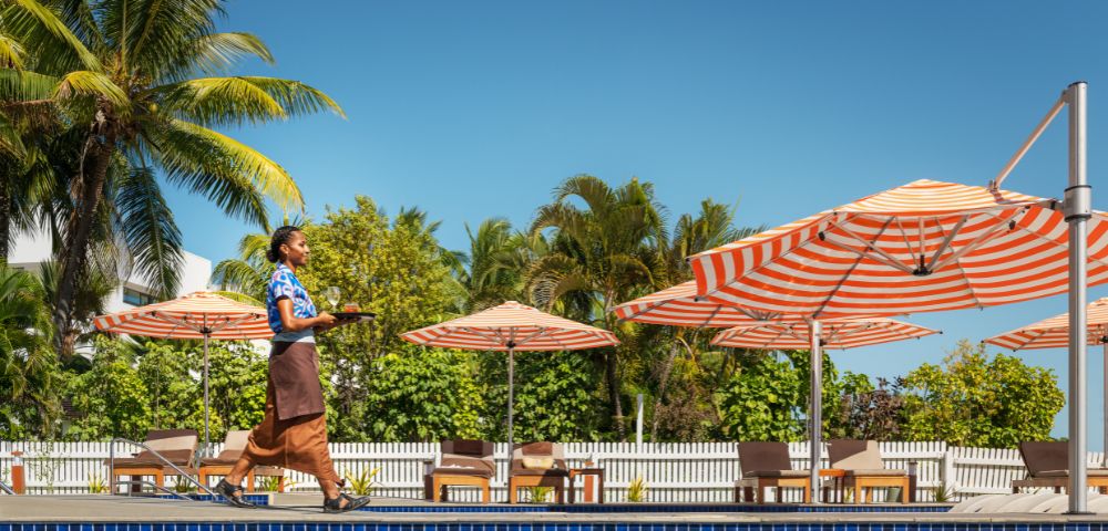Resort staff walking along a poolside deck carrying a tray, with orange-and-white striped umbrellas, lounge chairs, palm trees, and a clear blue sky in the background.