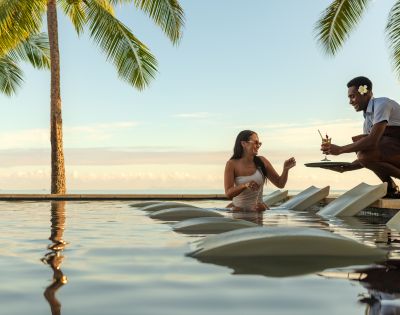 Resort guest seated in a pool on submerged lounge chairs receiving a drink from a staff member, with palm trees and ocean view in the background