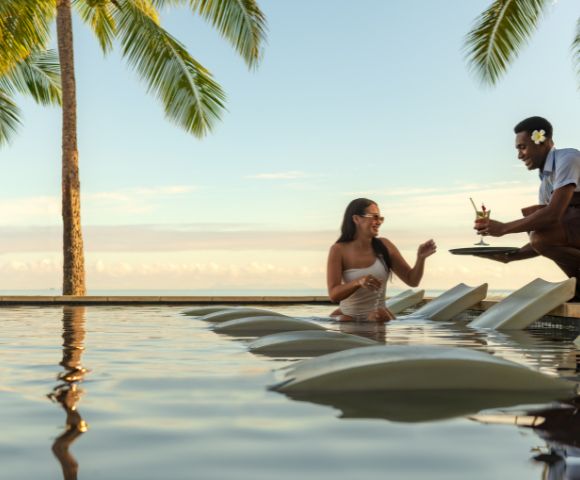 Resort guest seated in a pool on submerged lounge chairs receiving a drink from a staff member, with palm trees and ocean view in the background
