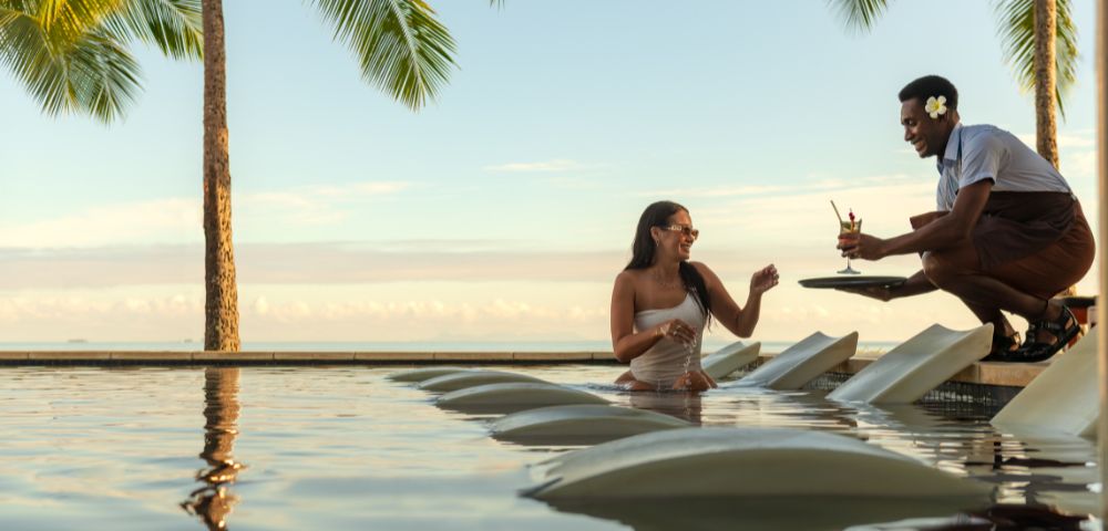 Resort guest seated in a pool on submerged lounge chairs receiving a drink from a staff member, with palm trees and ocean view in the background
