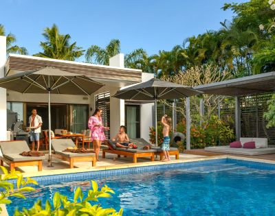 A family enjoying a sunny day by a pool at a modern villa. Adults relax on loungers under umbrellas, while children play nearby. Palm trees enhance the tropical setting.