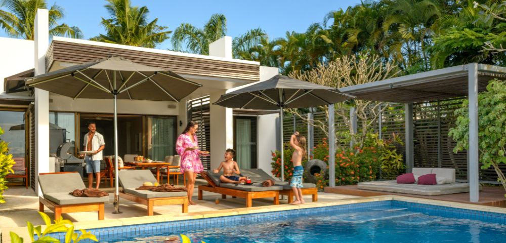 A family enjoying a sunny day by a pool at a modern villa. Adults relax on loungers under umbrellas, while children play nearby. Palm trees enhance the tropical setting.