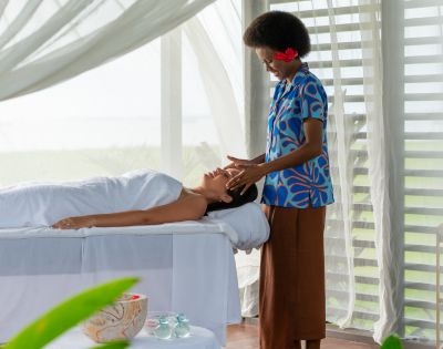 A serene spa scene with a relaxed woman lying on a massage table, covered with a white towel. A therapist in a blue patterned shirt gently massages her face. The room is airy with soft natural light, sheer curtains, and a peaceful ambiance.