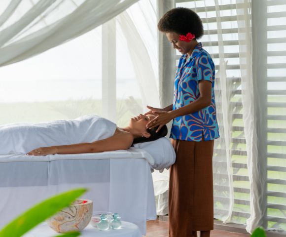 A serene spa scene with a relaxed woman lying on a massage table, covered with a white towel. A therapist in a blue patterned shirt gently massages her face. The room is airy with soft natural light, sheer curtains, and a peaceful ambiance.
