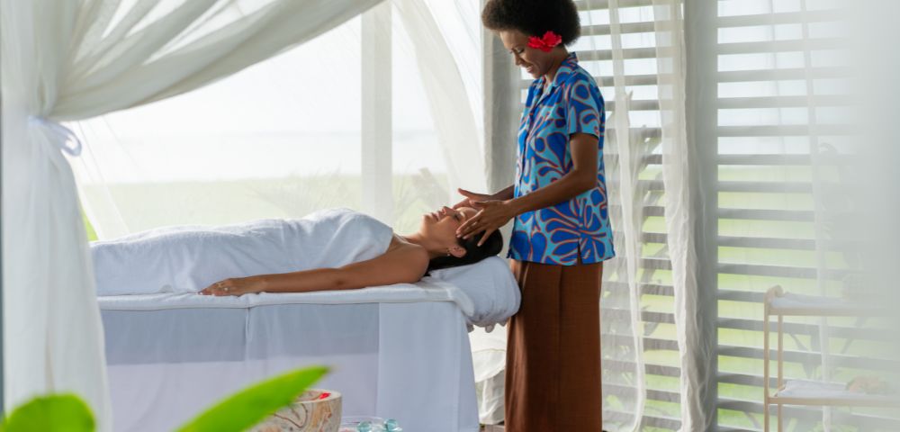A serene spa scene with a relaxed woman lying on a massage table, covered with a white towel. A therapist in a blue patterned shirt gently massages her face. The room is airy with soft natural light, sheer curtains, and a peaceful ambiance.