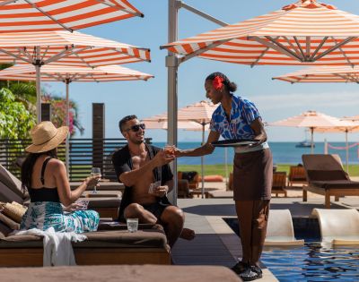 A couple relaxes on poolside lounges under orange-striped umbrellas, with a server handing drinks. The atmosphere is sunny and leisurely near the ocean.