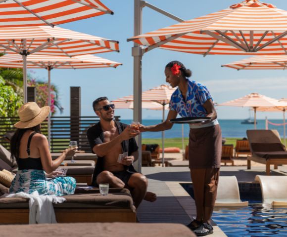 A couple relaxes on poolside lounges under orange-striped umbrellas, with a server handing drinks. The atmosphere is sunny and leisurely near the ocean.